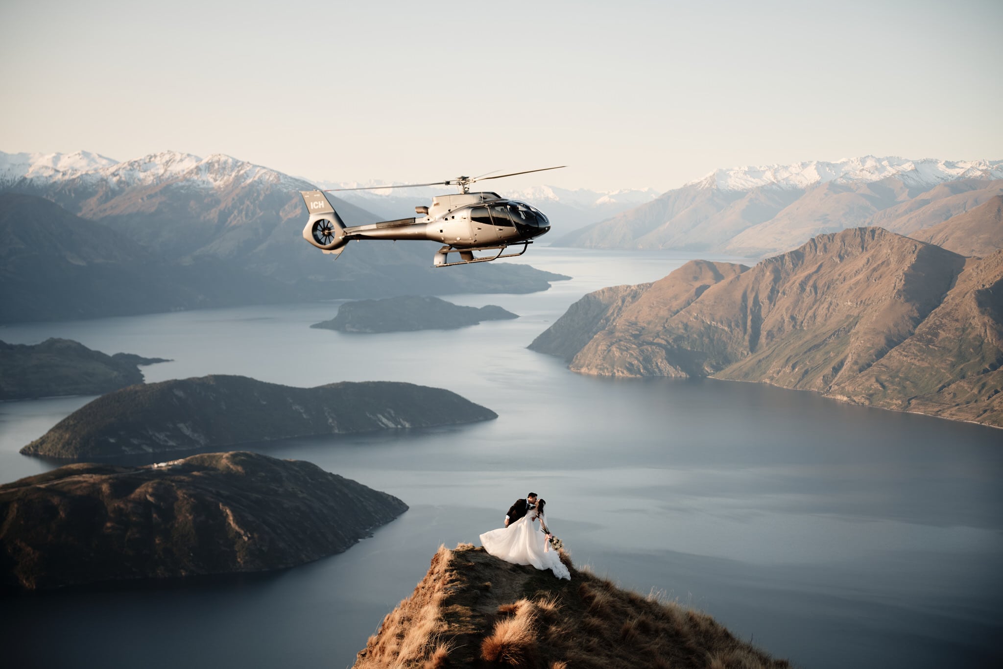クイーンズタウン　ニュージーランド　ウェディング　前撮り　挙式　結婚式　フォトグラファー | ヘリコプターを背景にクイーンズタウン山の頂上に立ち、ヘリウェディング＆エロープメントパッケージを楽しむ新郎新婦。